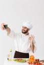 Image of caucasian cook man in uniform showing ok sign and taking selfie photo of food on smartphone at work isolated over white