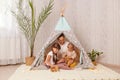 Image of Caucasian mother with her children sitting together teepee tent, mom playing with kids in play tent, mommy spending Royalty Free Stock Photo