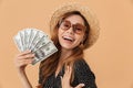 Image of charming european woman 20s wearing straw hat and sunglasses smiling and holding fan of dollar banknotes, isolated over