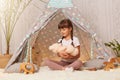 Image of charming little girl wearing white t shirt sitting on floor in wigwam and playing with teddy bear, holding toy in hands, Royalty Free Stock Photo