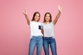 Image of charming two young twin sisters with beautiful smile with hands up showing peace gesture over pink background. Royalty Free Stock Photo