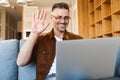 Image of cheerful adult man waving hand and working with laptop while