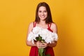 Image of cheerful energetic young lady wearing red dress, having pleasant facial expression, holding white peonies in both hands, Royalty Free Stock Photo