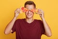 Image of cheerful handsome young boy holding two pieces of pizza in both hands, having beautiful smile, covering his eyes with