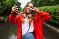 Image of cheerful young woman taking selfie on cellphone while walking through green park under rain Royalty Free Stock Photo