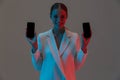 Image closeup of pretty woman 20s holding two mobile phones and showing black screens while standing under neon lights