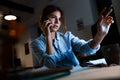 Young woman graphic designer using pc computer talking by mobile phone working at night in office. Royalty Free Stock Photo