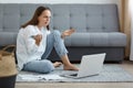 Image of confused helpless woman wearing white shirt and jeans sitting on floor near gray sofa, sitting with spreading hands