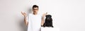 Image of confused man in glasses raising hands and shrugging complicated, standing near black pug dog over white Royalty Free Stock Photo