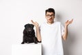 Image of confused man in glasses raising hands and shrugging complicated, standing near black pug dog over white Royalty Free Stock Photo
