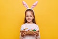 Image of cute pleased little girl wearing bunny ears holding Easter eggs in wicker basket isolated on yellow background, looking Royalty Free Stock Photo