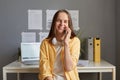 Image of delighted adorable young adult woman wearing yellow shirt sitting in office, talking on cell phone with, expressing Royalty Free Stock Photo