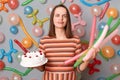 Image of disappointed irritated woman with brown hair wearing striped dress holding cake, looking at camera with desperate look,