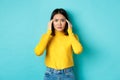 Image of distressed asian woman touching head and frowning, feeling headache, standing troubled against blue background