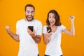 Image of ecstatic couple man and woman smiling while both using smartphones, isolated over yellow background