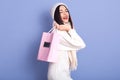 Image of emotional charismatic black haired young woman holding paperbag with purchase aback, wearing white sweater, hat and scarf