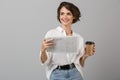 Emotional young business woman posing over grey wall background reading newspaper drinking coffee Royalty Free Stock Photo