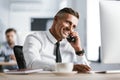 Image of european adult man 30s wearing white shirt and tie sitting at desk in office by computer, and speaking on smartphone Royalty Free Stock Photo