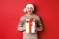 Image of excited handsome man receiving christmas gift, wearing santa hat and winter sweater, shouting for joy, holding