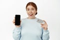 Image of excited smiling ginger girl shows her phone screen, empty mobile display and bank or discount credit card Royalty Free Stock Photo
