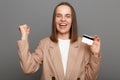 Image of excited smiling woman with brown hair wearing beige jacket posing isolated over gray background, winning lottery, showing