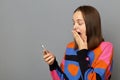 Image of excited woman with brown hair wearing jumper standing over grey background, holding cell phone, reading