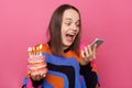 Image of excited woman holds tasty doughnuts with burning candles and smart phone, reading message with congratulations, wears