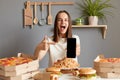 Image of extremely happy overjoyed woman with brown hair wearing white T-shirt sitting at table in kitchen, pointing at mobile Royalty Free Stock Photo