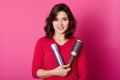 Image of female hairdresser holds comb and hairspay, wears red sweater, ready to make hairstyle her client, models over pink