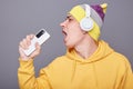Image of funny cheerful young adult man in beanie hat and yellow casual hoodie, listening his favorite playlist, holding smart