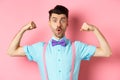 Image of funny guy with moustache and bow-tie, showing muscles, flexing biceps and looking surprised at camera, standing