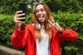 Image of funny young woman taking selfie on cellphone while walking through green park under rain Royalty Free Stock Photo