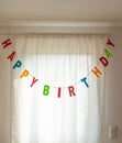 Image of garland with letters Happy Birthday hanging in front of a window with white curtain Royalty Free Stock Photo