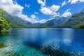 Serene blue lake with clear sky and white clouds above, encircled by lush greenery and towering mountains, creating a tranquil Royalty Free Stock Photo