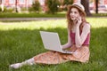 Image of good looking curly haired young female sitting on grass, wearing colorful skirt, red top and straw hat, relaxing in park Royalty Free Stock Photo