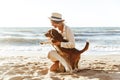 Image of gorgeous woman in straw hat hugging her brown dog, while walking by seaside