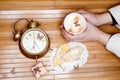 Image of hands holding cup of hot drink with pastries on plate, small gift box and alarm clock around Royalty Free Stock Photo