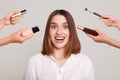 Image of the hands of several beauticians holding their respective equipment giving makeup to beautiful excited brunette girl