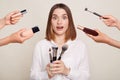 Image of the hands of several beauticians holding their respective equipment giving makeup to beautiful shocked astoinshed woman,