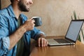 Image of handsome bearded man drinking coffee and using laptop while sitting at desk in cafe indoors Royalty Free Stock Photo