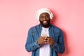 Image of handsome Black guy in headphones, listening music and using mobile phone, smiling at camera, pink background Royalty Free Stock Photo