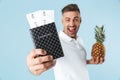 Handsome excited happy adult man posing  over blue wall background holding passport and pineapple Royalty Free Stock Photo