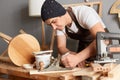 Image of handsome guy carpenter wearing apron and cap working with plane on wooden workplace background, joiner making wooden