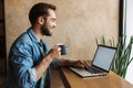 Image of handsome happy man drinking coffee and using laptop while sitting at desk in cafe indoors Royalty Free Stock Photo