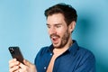 Image of handsome young man feeling happy reading good news on mobile phone, standing cheerful against blue background