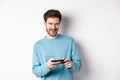 Image of handsome young man playing mobile video game, holding smartphone horizontally and smiling at camera pleased Royalty Free Stock Photo