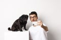 Image of handsome young man taking selfie with cute black dog on smartphone, posing with pug over white background