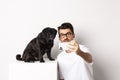 Image of handsome young man taking selfie with cute black dog on smartphone, posing with pug over white background