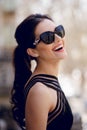 Smiling beautiful brunette model, in elegant black dress and chic sunglasses, ponytail, posing outside. Royalty Free Stock Photo