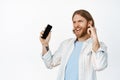 Image of happy blond guy dancing with earphones, listening music in headphones, holding mobile phone and smiling pleased Royalty Free Stock Photo